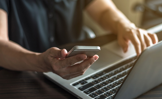 One hand holding smartphone while other hand types on laptop keyboard
